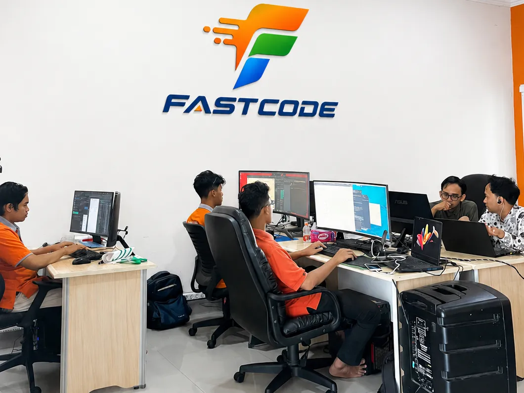 Lima orang tim FASTCODE bekerja di meja masing-masing di ruang kerja kantor, di bawah lambang dan nama FASTCODE yang terpasang di dinding.